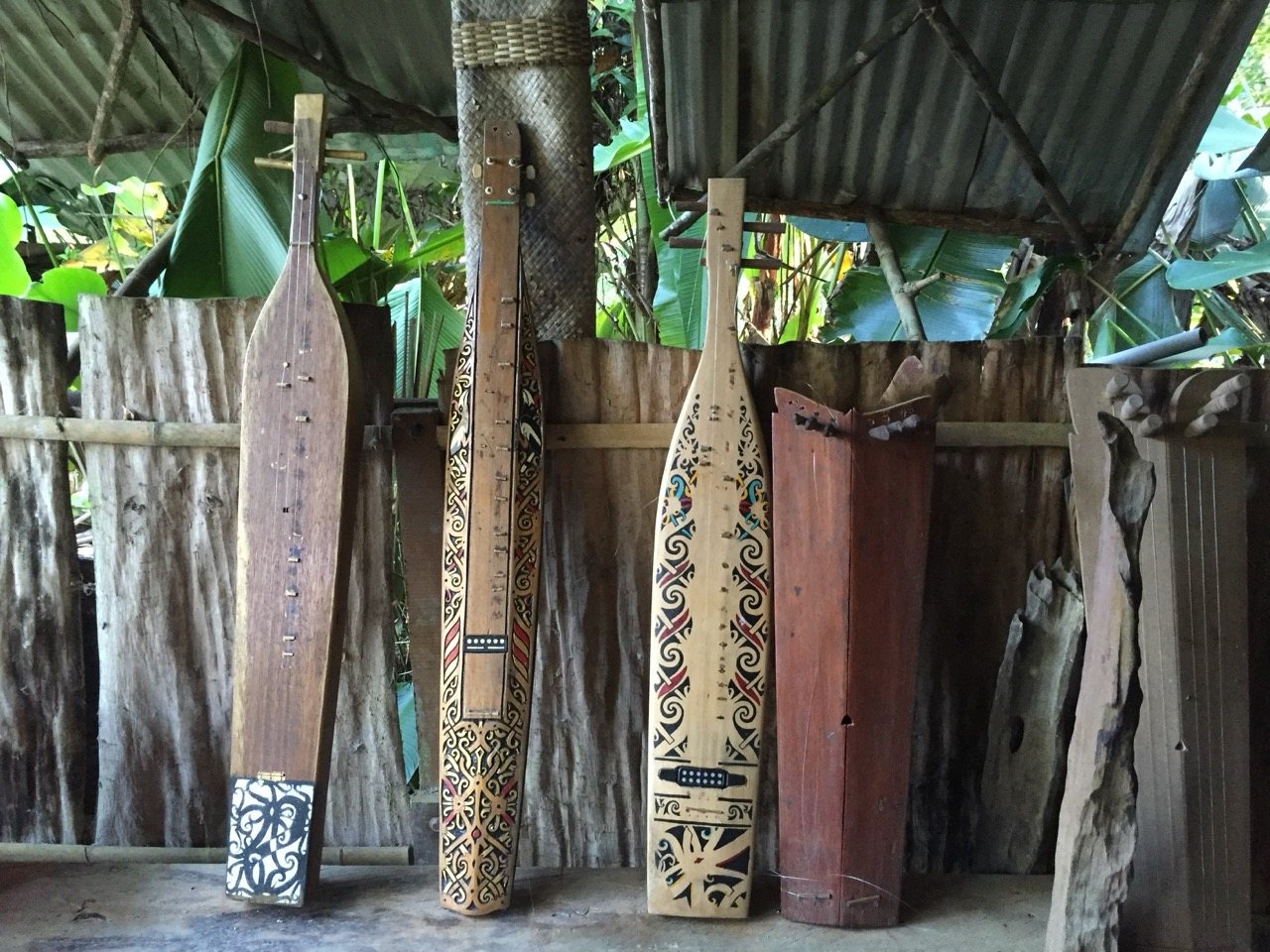Four sapes of different sizes and decorated bodies leaning against a bamboo and corrugated wall, their carved surfaces facing outward.