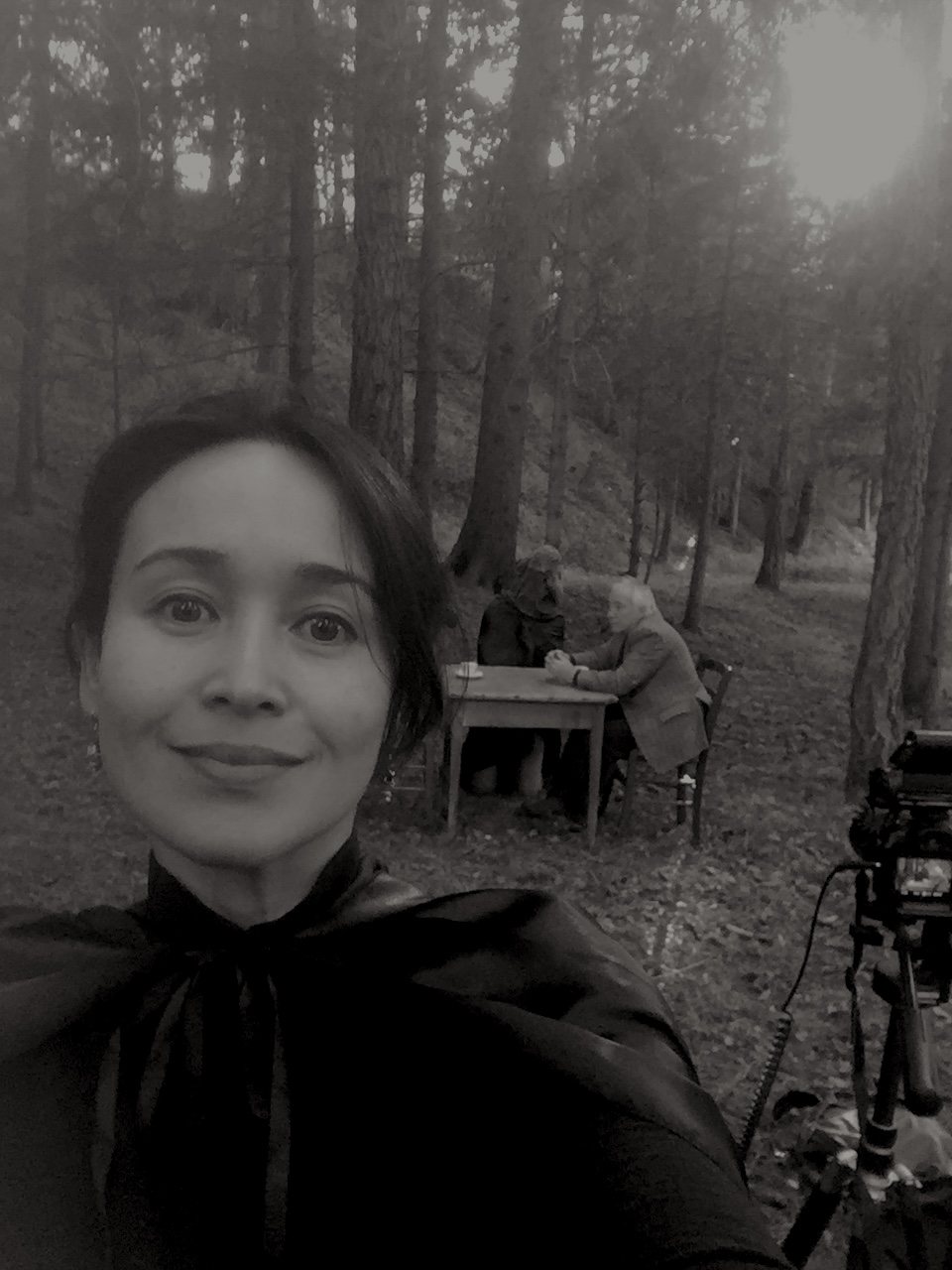 A self-portrait of the filmmaker wearing the black cloak, smiling at the camera, the artist and the seated tripod visible in the forest behind her.